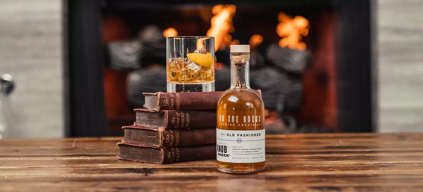 Old fashioned bottle next to a stack of old books with a filled glass on top with fireplace in background.