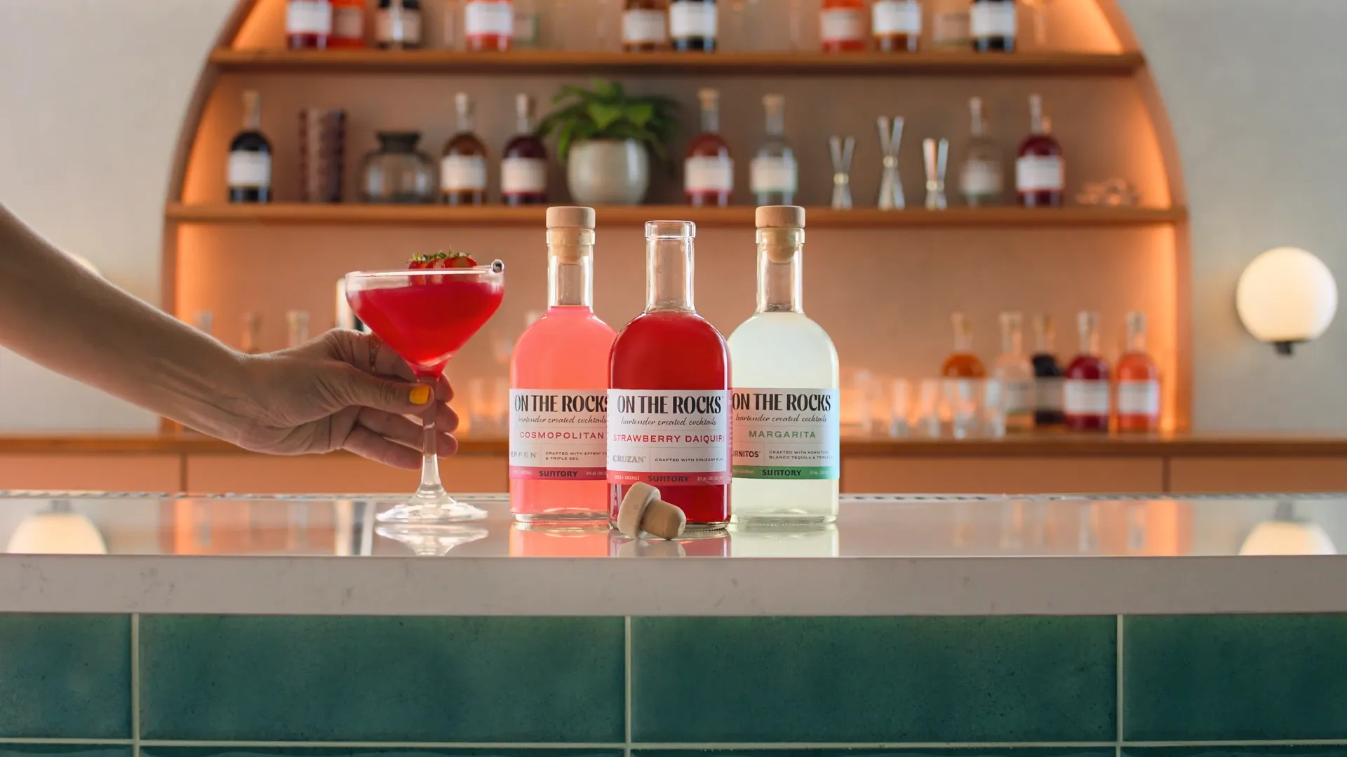 A hand holds a vibrant red cocktail on a bar, next to three bottles of ready-to-drink cocktails: Cosmopolitan, Strawberry Daiquiri, and Margarita.